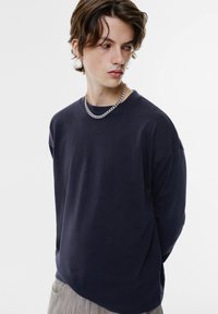 Dunkelblaues Langarmshirt aus Baumwolle mit Rundhalsausschnitt, lockerer Passform, überschnittenen Schultern und glatter Textur. Akzent mit einer silbernen Kettenhalskette.