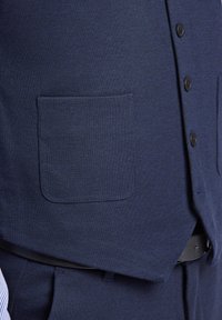 Marineblauer Weste mit strukturierter Oberfläche, ausgestattet mit einer einzelnen Brusttasche, fünf schwarzen Knöpfen und einer flachen Naht am unteren Rand.