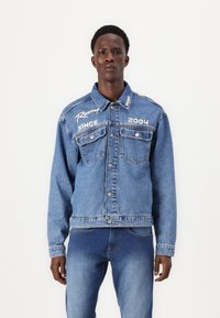 Chaqueta de mezclilla azul con cierre de botones, dos bolsillos en el pecho y texto bordado en blanco que dice "Compitiendo desde 2004". Tejido texturizado con un corte holgado.