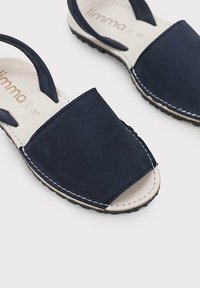 Sandalias de ante azul marino con la puntera descubierta, una tira delantera gruesa y dos tiras traseras. Presentan costuras beige y una suela negra texturizada.
