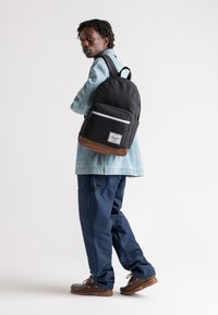 Zaino nero con base marrone, tasca frontale con zip e dettagli bianchi. Indossato da una persona in giacca azzurra chiara e pantaloni blu navy.
