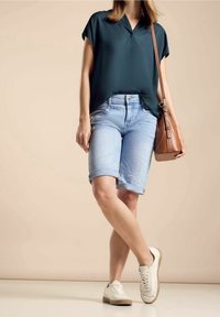 Kvinna som bär en mörkgrön blus med korta ärmar, ljusblå uppvikta jeansshorts, vita sneakers och bär en brun läderväska över axeln.