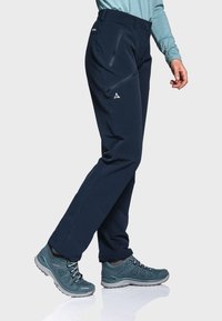 Navy Softshell-Hosen mit Reißverschlusstaschen, entworfen für den Außeneinsatz, mit einem elastischen Bund und schmal zulaufenden Beinen. Begleitet von tealfarbenen Wanderschuhen.