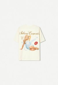Cremefarvet T-shirt med et grafisk print af en kvinde i en blå kjole, ledsaget af tekst og et rødt læbeprint som accent.