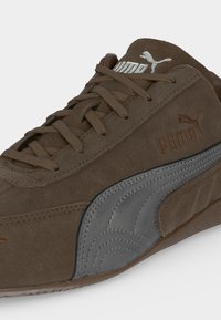 Кафяв велурен маратонка Puma с кафяви връзки и сив детайл с ивица Puma отстрани, както и лого на Puma на езика и отстрани.