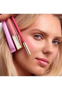 Produkty do ust Estée Lauder w tubkach—różowy, brzoskwiniowy i fioletowy—umieszczone blisko twarzy modelki z delikatnym makijażem i blond włosami. Widoczne złote akcenty.