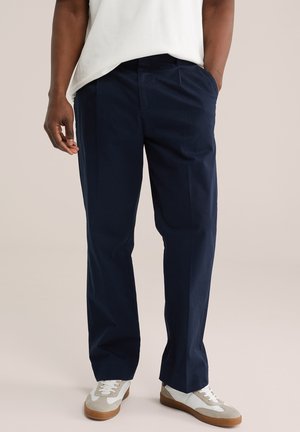 Muž v tmavomodrých nohaviciach s priamym strihom, bielo-béžových teniskách a bielej košeli, s jednou rukou v vrecku.