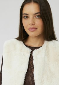 Jeune femme aux longs cheveux bruns portant un gilet en fausse fourrure couleur crème par-dessus un haut en dentelle marron foncé, faisant légèrement face à gauche avec une expression neutre.