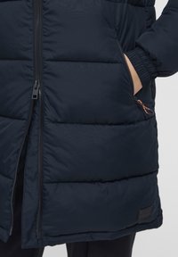 Puffer jacket in navy, featuring a zip closure, side pockets, and elastic cuffs. Black hardware and brand label on the hem. Smooth texture.
