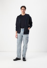 Felpa blu navy con zip e maniche a fantasia, t-shirt nera sotto, pantaloni cargo blu chiaro con tasche e scarpe nere.