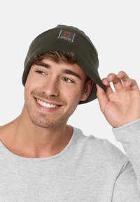 Dark green ribbed beanie with a rectangular patch featuring the text "JEANSCODE." Model wears a light gray sweater.