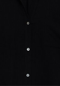 Gros plan sur une chemise en tissu noir montrant trois boutons blancs brillants et une légère silhouette d'une poche du côté droit.