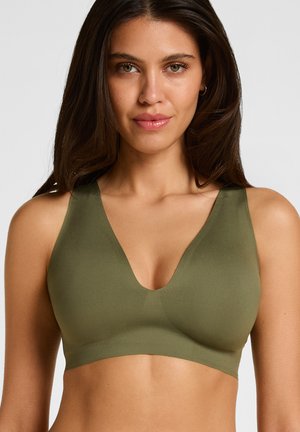 Woman with long dark hair wearing sleeveless olive green bra top, facing forward against a plain light background.