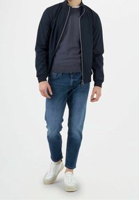 Giacca bomber marina con cerniera frontale, maglione grigio, jeans blu e sneakers bianche con accenti beige. Tessuto liscio e vestibilità casual.