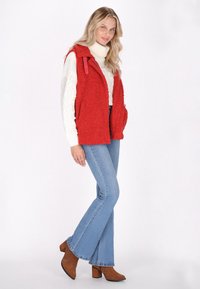 Rote, flauschige Weste mit Kragen, Seitentaschen und Druckknöpfen, getragen über einem weißen Pullover und kombiniert mit hellblauen, weit geschnittenen Jeans und braunen Stiefeletten mit Absatz.