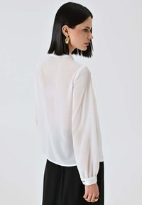 Blusa bianca a maniche lunghe con una texture morbida e trasparente, con bottoni decorativi ai polsini, abbinata a una gonna nera.