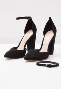 Paire de chaussures à talons hauts en daim noir avec bouts pointus, talons carrés, brides à la cheville et une sangle noire séparée avec une boucle dorée.