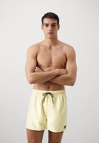 Žlté plavkové šortky s elastickým pásom a čiernym šnúrkou. Hladká látka v jednoliatej farbe. Model pózuje so založenými rukami.