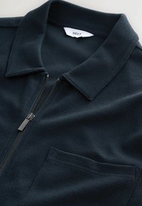 Tmavomodré polo tričko na zips s textúrovanou látkou, krátkym golierom a jedným náprsím vreckom; obsahuje kovový zips a štítok značky vo vnútri.