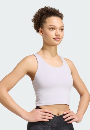 Joven mujer con cabello rizado, con un top corto de color púrpura claro y leggings negros con estampado, de pie con las manos en las caderas, mirando hacia la derecha.