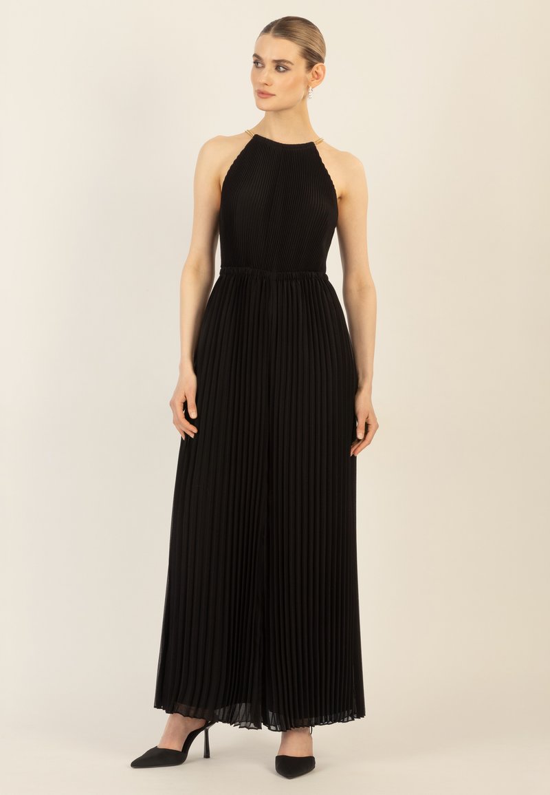 Mustapleated jumpsuit, jossa on halterneck-olkaimet, kultaiset yksityiskohdat hartioissa ja virtaava siluetti. Mukana on mustat korkokengät.