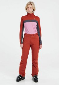 Pettorina da sci rossa e rosa con colletto alto, caratterizzata da accenti grigi. Abbinata a pantaloni da sci rossi. Stivali da sci neri con fibbie metalliche.