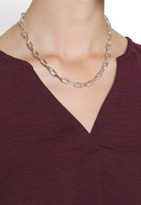 Collier en chaîne en argent avec des maillons en forme d'oval, associé à un haut en v bourgogne. Le haut a un tissu texturé avec des coutures subtiles.