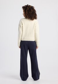 Maglione di colore crema, con texture, abbinato a pantaloni larghi blu navy con strisce bianche ai lati, visto da dietro.