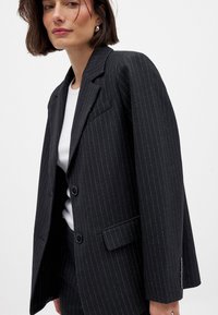 Blazer noir à rayures avec une coupe ajustée, fermeture à un bouton, revers crantés et deux poches avant. Tissu texturé avec un subtil éclat.