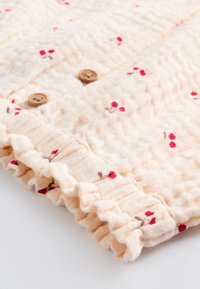 Tissu crème texturé avec de petits motifs floraux rouges ; comprend des boutons en bois et un ourlet à volants pour plus de détails.