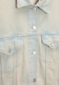 Chaquetón de mezclilla azul claro con botones de presión plateados, dos bolsillos en el pecho y un clásico cuello puntiagudo. Textura desgastada y detalles de costura.