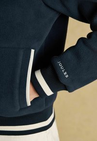 Klahvide fleece-jakk valgete aktsentidega; ribidega varrukad ja vöökoht; varrukal on tikandiga "JOULES".