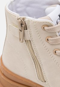 Beżowe trampki typu high-top z bocznym zamkiem, beżowe sznurowadła przeprowadzone przez metalowe oczka, biała wyściółka kołnierza i brązowa gumowa podeszwa.