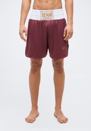 Ανδρικός κορμός και πόδια που φοράνε μπορντό σορτς πυγμαχίας Venum με λευκή μέση, όρθιος ξυπόλητος μπροστά σε απλό φόντο.