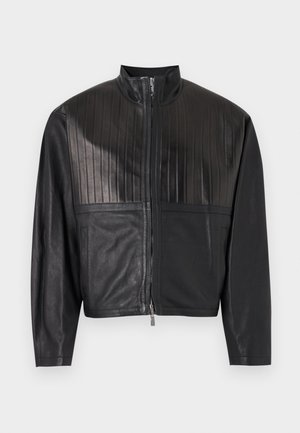 Man wearing black leather jacket with vertical pleats, olive green shirt, black leather pants, and black shoes standing against white wall.