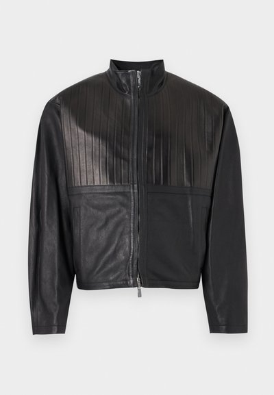 Μαύρο μπουφάν από συνθετικό δέρμα με κάθετες ραφές στο στήθος, εμπρόσθιο φερμουάρ και δύο πλαϊνές τσέπες, παρουσιασμένο σε απλό φόντο.