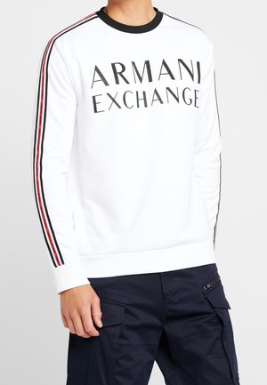 Sweatshirt blanc à manches longues Armani Exchange avec des détails rayés noirs et rouges sur les manches, porté avec un pantalon cargo foncé.