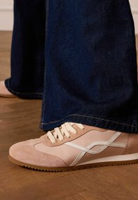 Sneaker bassa in suede rosa e materiale sintetico con dettagli bianchi e lacci color crema. Presentata con pantaloni in denim scuri a gamba larga e pavimento in legno.