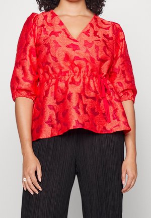 Woman wearing a red floral textured blouse with 3/4 puffed sleeves and a waist tie, paired with black pleated wide-leg pants.