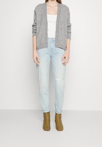 Cárdigan de punto gris, camiseta sin mangas blanca, jeans de mezclilla azul claro con detalles desgastados y botas de tobillo oliva con punta afilada.