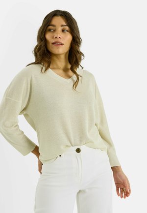 Suéter de cuello en V color beige claro con mangas de tres cuartos, hecho de una tela suave y brillante. Combinado con pantalones blancos de tiro alto y detalles de herrajes visibles.