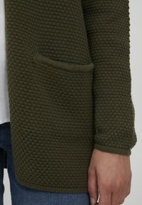 Pletený kardigan ve tmavě olivové barvě s texturovaným vaflovým vzorem. Má kapsu a žebrované manžety, s klasickým otevřeným designem v přední části.
