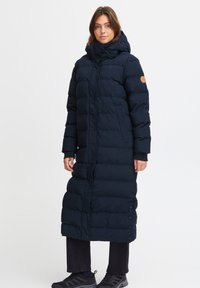 Lang, marineblå pufferjakke med hette, med knappelukking foran, quiltet design og elastiske mansjetter. Akzentuert med en logo på ermet.