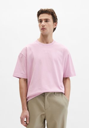 Man draagt een lichtroze oversized T-shirt en beige broek, staand met één hand in de zak tegen een effen witte achtergrond.