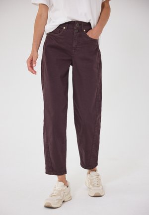 Persona che indossa pantaloni a vita alta color viola scuro con gamba dritta e bottoni frontali, e scarpe da ginnastica bianche, in piedi davanti a uno sfondo semplice.