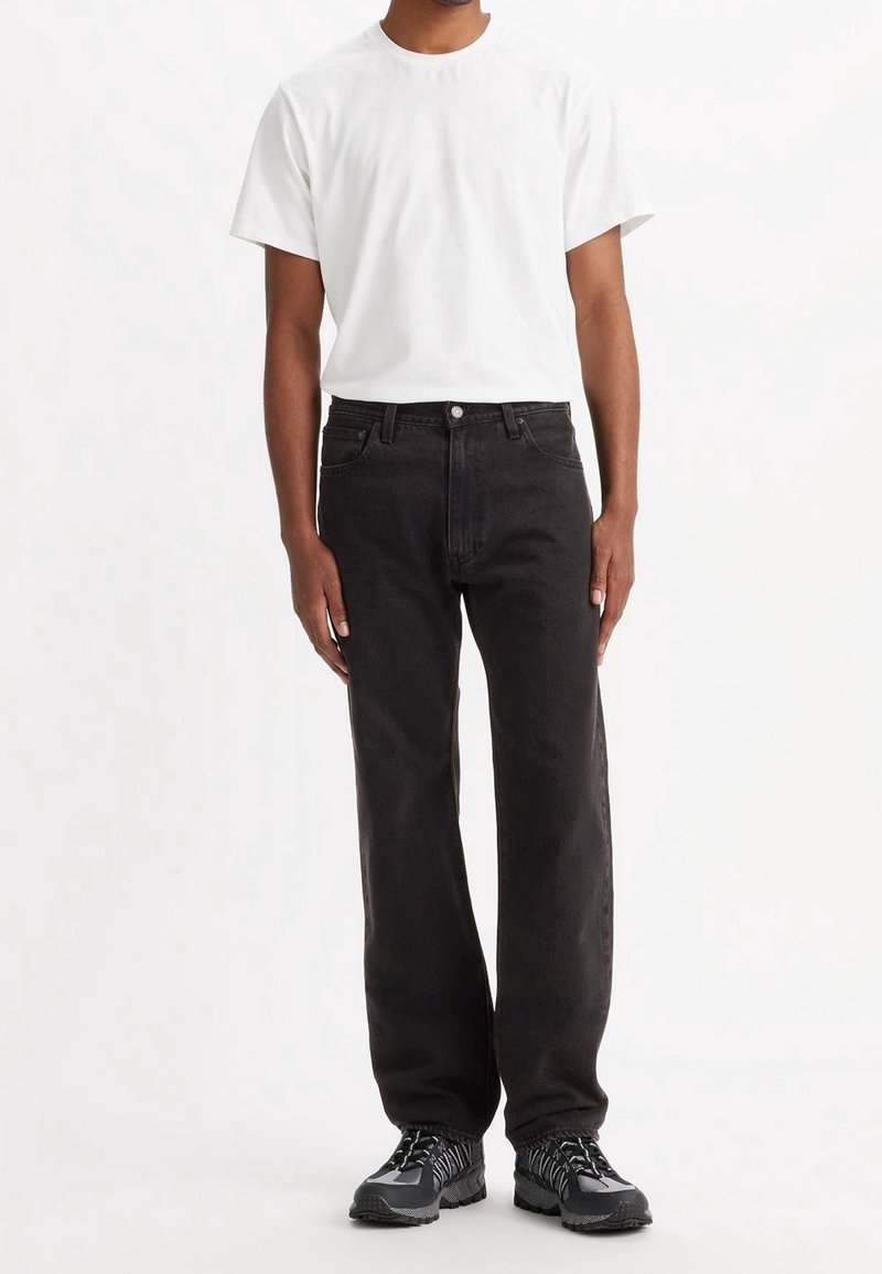 Personne portant un t-shirt blanc uni à manches courtes, un jean noir droit et des baskets gris et noires, debout, les bras le long du corps.