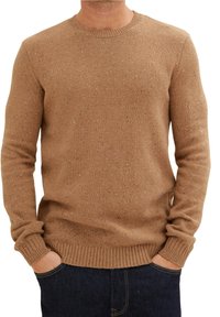 Mann in einem braun gesprenkelten Strickpullover mit gerippten Bündchen und Saum, die Hände in den Taschen einer dunkelblauen Jeans.