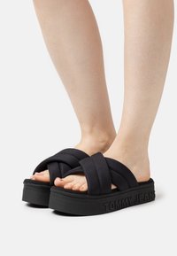 Schwarze Plateausandalen mit gewundenem Gewebe-Obermaterial, ausgestattet mit einer strukturierten Gummisohle und geprägtem "TOMMY JEANS"-Logo an der Seite.