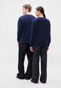 Tengerészkék pulóverek bordázott mandzsettával és szegéllyel, sötét farmernadrággal viselve. Mindkét ruhadarab laza szabású és minimális díszítéssel rendelkezik.