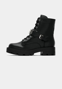 Botas de cuero negras hasta el tobillo con suela gruesa, acentos de metal y una correa con hebilla. Presentan ojales y cordones para el cierre.